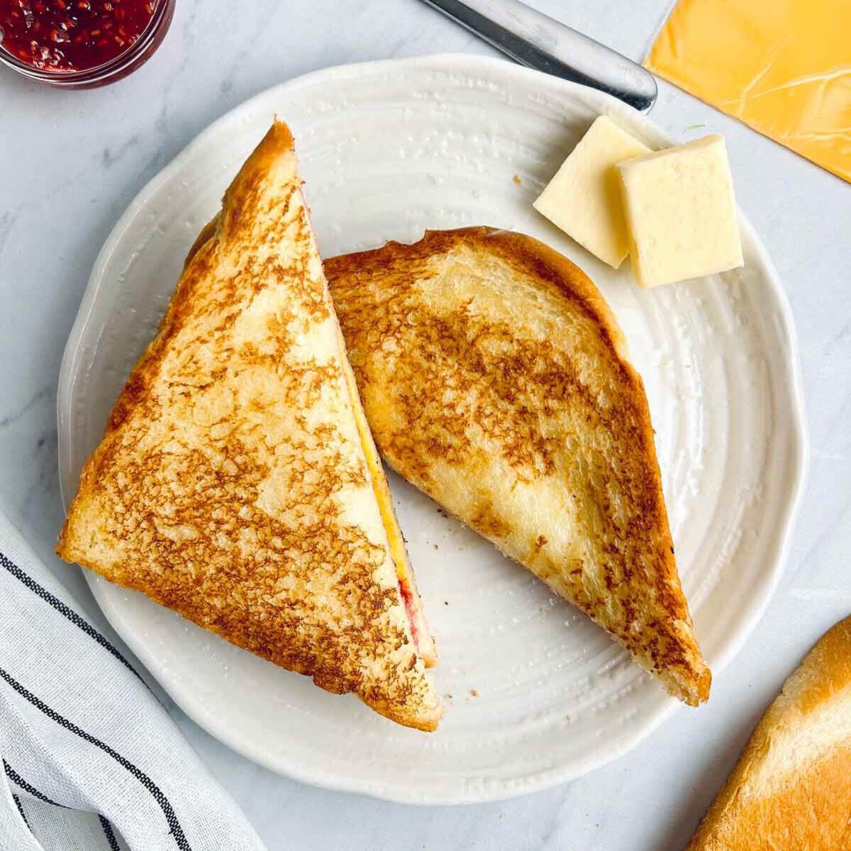 Grilled jam & cheese sandwich on a white plate with two cubes of butter, with red jam in a ramekin in the top left hand corner