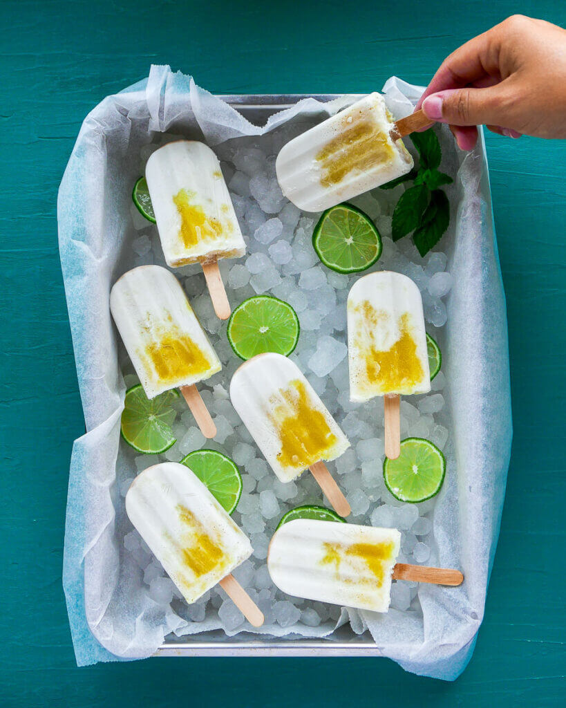 Coconut mango popsicles atop ice atop parchment paper, decorated with lime slices, on a turquoise wood background