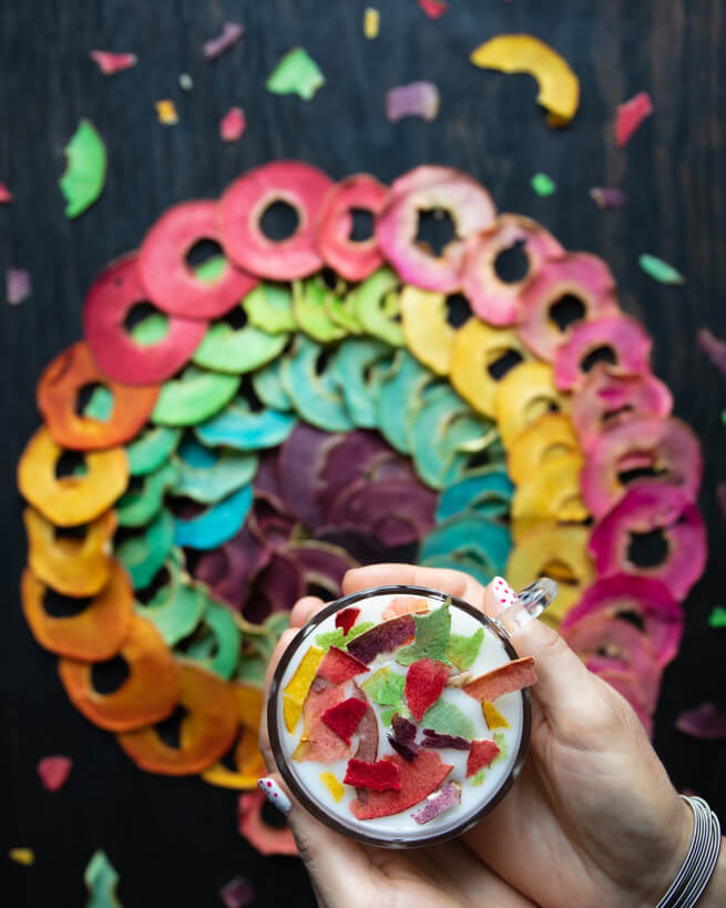 Rainbow-colored dried apple slices arranged in a circle. Two hands are holding a cup of white yogurt topped with colorful dried apple slices