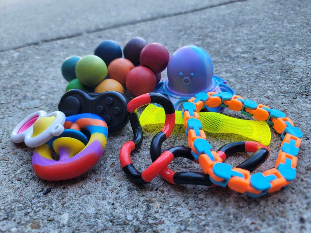 8 fidget and sensory tools on pavement; including: a blue and orange clicking snake fidget, a red and black wavy fidget, a circle silicone pop fidget, a black mini game controller fidget, a silicone octopus fidget, a white plastic duo pop fidget with yellow and pink circles, a yellow marble fidget, and a rainbow-colored fidget with wooden balls